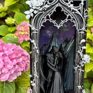 Könnte beinhalten: Ein rechteckiger schwarzer Schattenkastenrahmen mit einem verzierten silbernen gotischen Torbogen, der mit skulptierten Rosen verziert ist. Im Inneren steht eine schwarze 3D-gedruckte Figur von zwei sich umarmenden Personen vor einem violetten Hintergrund.