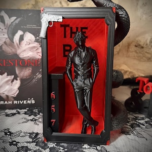 May include: A black rectangular display frame with a red interior featuring a 3D figure of a person standing with hands in pockets. A black snake coils around the top, a silver pistol is attached to the corner, and the numbers 6, 5, 7 are stacked vertically.