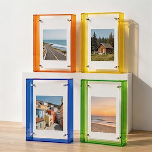 Könnte beinhalten: Vier rechteckige Acryl-Fotorahmen in Orange, Gelb, Blau und Grün. Jeder Rahmen hat einen dicken, transparenten Rand und vier silberfarbene Metallschrauben an den Ecken, die ein weißes Passepartout und ein Foto im Inneren halten.