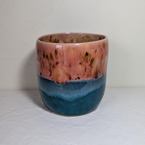 Peut inclure: Un gobelet en céramique fait à la main avec un dégradé de couleurs unique. La moitié supérieure est un mélange de rose et de marron, passant à un bleu sarcelle profond en bas. La tasse a une finition lisse et brillante.