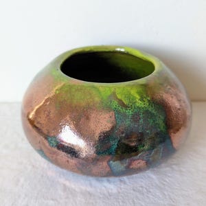 Könnte beinhalten: Eine handgefertigte Keramikvase mit runder Form und breiter Öffnung. Die Vase hat eine Mischung aus Kupfer-, Grün- und Türkistönen, die ein einzigartiges, künstlerisches Aussehen erzeugt. Der Innenraum ist dunkel und kontrastiert mit der glänzenden Oberfläche.
