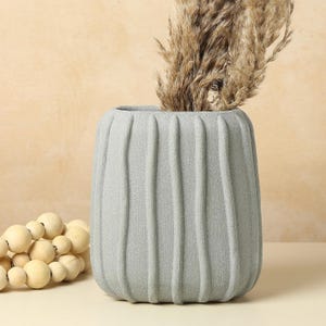 Puede incluir: Un jarrón gris texturizado con ranuras verticales, que contiene hierba ornamental seca. El jarrón tiene forma redondeada y se apoya sobre una superficie clara. Se ven cuentas decorativas de madera en la esquina inferior izquierda.