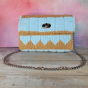 May include: A handmade, light blue and tan crochet handbag with a silver chain strap. The bag features a textured design with a silver clasp closure. The bag's design includes a zig-zag pattern along the bottom edge.