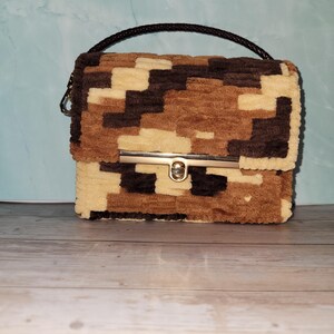 May include: A rectangular handbag with a camouflage pattern in shades of brown and beige. The bag features a braided black handle and a gold-colored clasp. The bag is made of a soft, textured material.