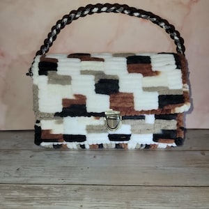 May include: A handmade rectangular handbag with a woven handle. The bag has a patchwork design in white, brown, black, and grey. It features a metal clasp closure and a braided handle in brown and white.