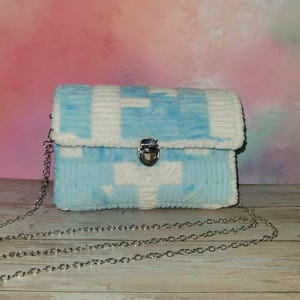 Peut inclure: Sac bandoulière bleu clair et blanc avec une chaîne argentée. Le sac est fait d'un tissu texturé avec un motif de croix blanches sur fond bleu. Il possède une fermeture à fermoir argenté.