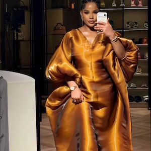 May include: A woman in a full-length, flowing, golden-brown dress with wide sleeves, posing in front of a mirror. The dress has a V-neck and a shiny, satin-like texture. The background includes a display of handbags and shoes.