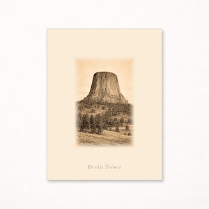 May include: A rectangular print on cream-colored paper featuring a sepia-toned illustration of Devils Tower rising above a forest and grassy landscape. The text "Devils Tower" is printed in a simple font at the bottom center of the image area.
