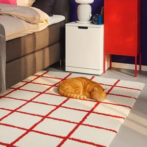 Puede incluir: Interior de dormitorio con una alfombra blanca con un diseño de cuadrícula roja. Un gato atigrado naranja está acurrucado durmiendo sobre la alfombra. Una cama con cabecero gris y ropa de cama blanca está al fondo, junto a una mesita de noche blanca y un armario rojo.