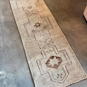 May include: A long, rectangular runner rug featuring a faded beige and cream base color with dark brown geometric patterns. The design includes repeating star-like motifs and angular shapes arranged in a central column along the length of the rug.