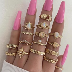May include: A hand displaying a collection of gold rings with various designs, including opals and intricate patterns. The rings are worn on fingers with long, pink nails. The rings are made of gold.