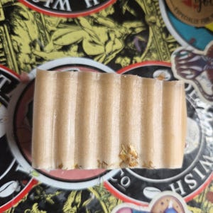 May include: A rectangular bar of handmade soap with a light brown color and a ribbed texture. The soap has small, visible particles and is likely scented. The soap is displayed on a surface covered with colorful stickers.
