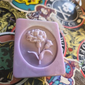 May include: A rectangular bar of lavender-colored soap with a floral design. The soap features a raised image of a flower with petals, leaves, and buds. The soap is set against a background of colorful stickers.