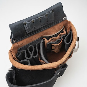 Op de afbeelding: Open zicht op een leren gereedschapsriem met meerdere zakken en compartimenten. De riem heeft een combinatie van zwart en bruin leer, met een lichtere bruine binnenkant. De riem heeft een metalen clip en klinknagels ter versteviging.