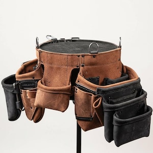 May include: A brown leather tool belt with multiple pockets and pouches. The belt has a circular top with metal rings and a black interior. Black and brown pockets are attached to the belt, designed for various tools. The belt is displayed on a black stand.