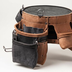 May include: A brown leather tool belt with multiple pockets and a black suede pouch. The belt has a circular top and a metal D-ring. Rivets and stitching detail the construction. A metal hook is attached to the side.