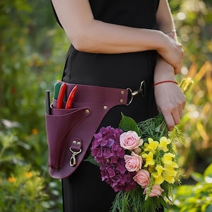 Könnte beinhalten: Ein burgunderfarbener Leder-Gartengerätegürtel mit einer Tasche, die grüne und rote Scheren und einen Stift enthält. Der Gürtel wird um die Taille getragen und hat einen goldfarbenen Verschluss. Ein Blumenstrauß wird in der Nähe gehalten.