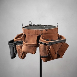 May include: A brown leather tool belt with multiple pockets and loops. The belt has a black interior and trim, with metal D-rings and rivets. The tool belt is designed for construction or carpentry work.
