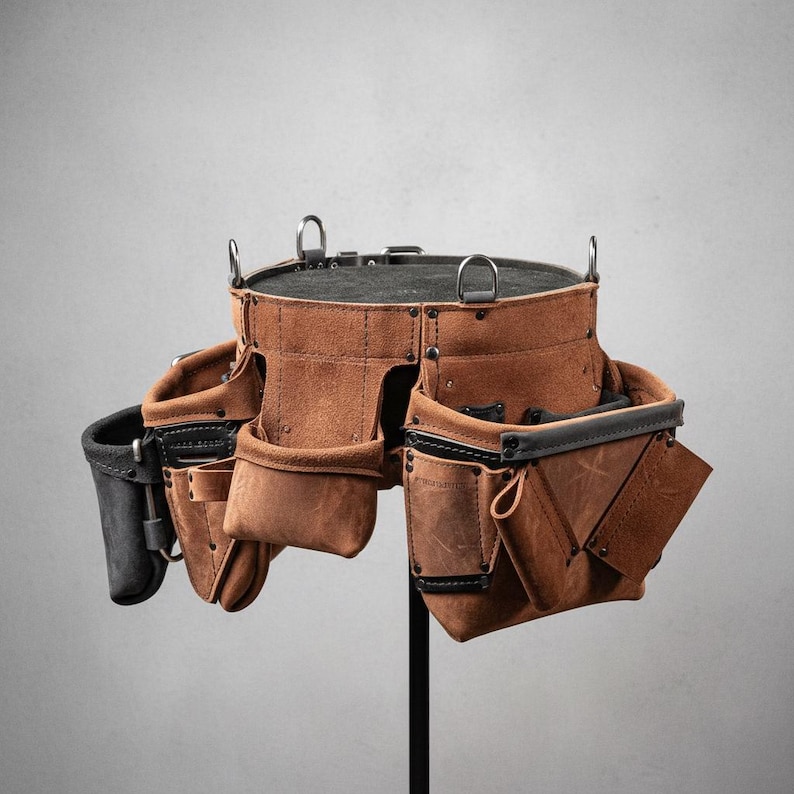 May include: A brown leather tool belt with multiple pockets and loops. The belt has a black interior and trim, with metal D-rings and rivets. The tool belt is designed for construction or carpentry work.