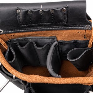 May include: A black and tan leather tool pouch featuring multiple open compartments and pockets for storage. The interior is lined with a natural tan suede texture, while the exterior is constructed from smooth black leather with metal rivets.