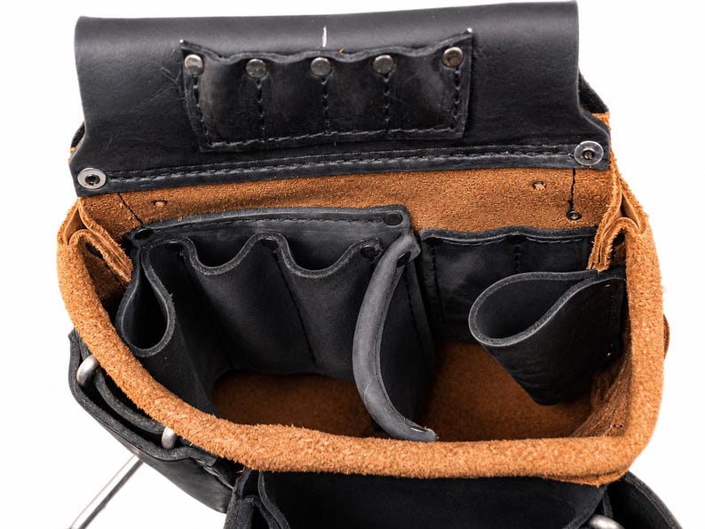 May include: A black and tan leather tool pouch featuring multiple open compartments and pockets for storage. The interior is lined with a natural tan suede texture, while the exterior is constructed from smooth black leather with metal rivets.
