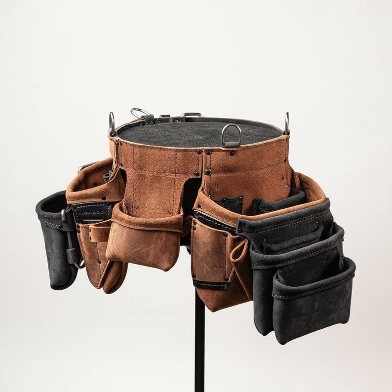 May include: A brown leather tool belt with multiple pockets and pouches. The belt has a circular top with metal rings and a black interior. Black and brown pockets are attached to the belt, designed for various tools. The belt is displayed on a black stand.
