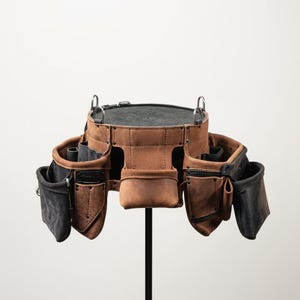 May include: A brown leather tool belt with multiple pockets and loops. The belt has a dark gray interior and is suspended by metal rings. The leather has a distressed appearance, suggesting durability and use.