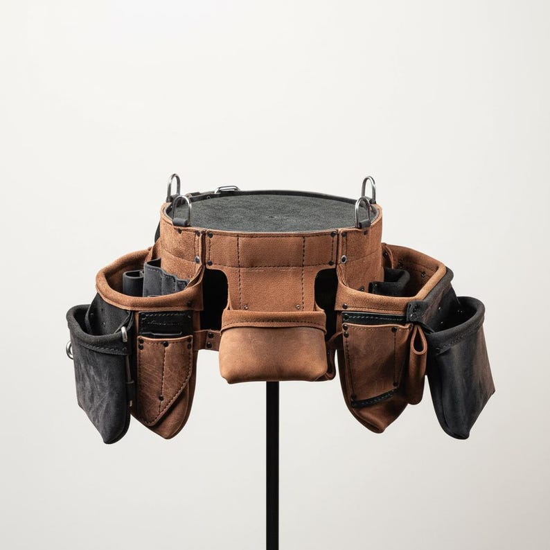 May include: A brown leather tool belt with multiple pockets and loops. The belt has a dark gray interior and is suspended by metal rings. The leather has a distressed appearance, suggesting durability and use.
