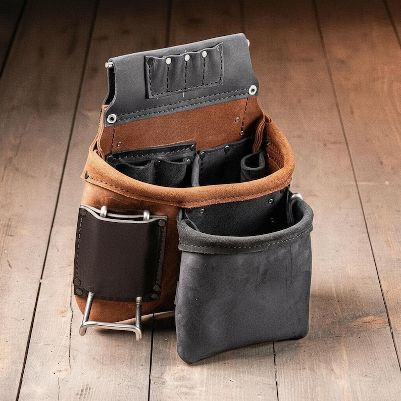 May include: A brown and black leather tool pouch featuring multiple compartments, a metal hammer loop, and reinforced stitching. The design includes a large open pocket, smaller interior slots for tools, and a sturdy belt attachment point.