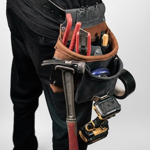 May include: A brown and black leather tool belt worn on the waist, featuring multiple pockets and loops. It holds a metal hammer with a red handle, a utility knife with a red casing, a yellow-handled tool, a black power drill, and a roll of black tape.