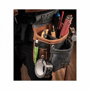 May include: A brown and black leather tool pouch attached to a belt, featuring multiple pockets and loops. It contains a red utility knife, a yellow-handled tool, a green marker, and a roll of black tape hanging from a metal hook on the front.