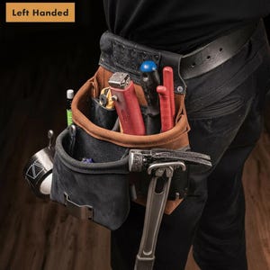 May include: A left-handed tool belt made of brown and black leather, featuring multiple pockets and loops. It holds a red utility knife, a black-handled screwdriver, a red tool, a silver hammer, and a roll of tape with a geometric pattern.