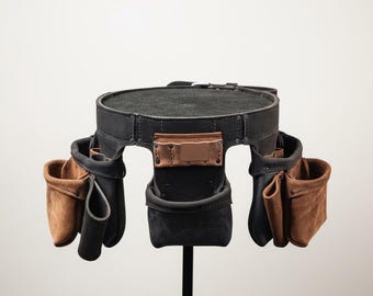 May include: A black leather tool belt featuring multiple brown leather pouches and pockets attached to the sides and front. The belt is constructed with reinforced metal rivets and includes a central rectangular leather loop for tool storage.