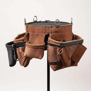 May include: A brown leather tool belt with multiple pockets and loops. The belt has a black accent on the edges and is designed to hold various tools. It is displayed on a black stand against a white background.