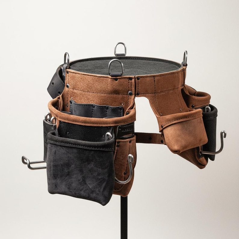 May include: A brown leather tool belt with multiple pockets and loops. The belt has a circular top with a black interior and is supported by a metal stand. The pockets are various sizes and include black and brown leather.