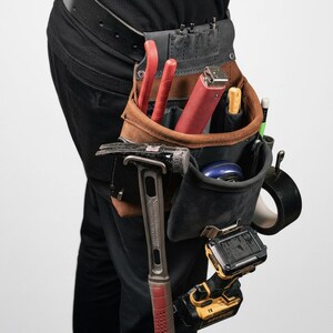 May include: A brown and black leather tool pouch attached to a black belt, containing a hammer, a red handled tool, a yellow handled tool, a blue circular object, a roll of black tape, and a yellow and black cordless power drill.