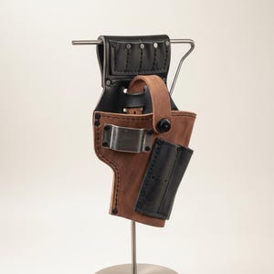 Puede incluir: Una funda de cuero marrón y negro con un clip metálico y remaches. La funda tiene un lazo y una pequeña bolsa adjunta. Se exhibe en un soporte metálico.