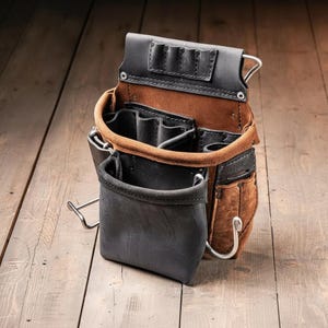 May include: A tool pouch constructed from brown and black leather, featuring multiple compartments, slots for small tools, and metal hooks. The design includes a large front pocket, a top section with three narrow slots, and side attachments for hardware.