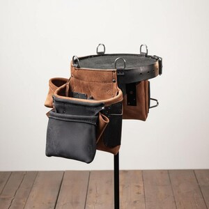 May include: A brown and black leather tool belt with multiple pockets and metal ring attachments. The belt is designed for carrying tools and is displayed on a black stand against a neutral background. The leather has a textured appearance.