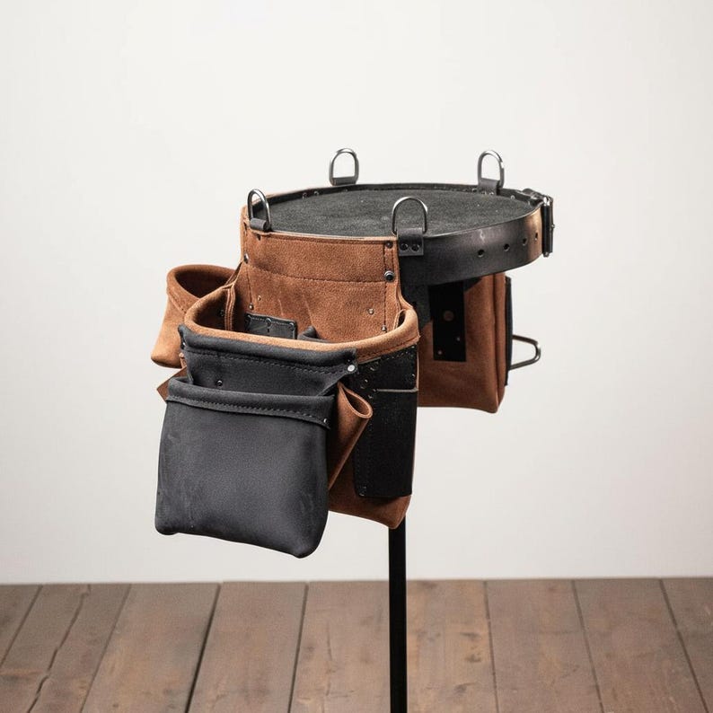 May include: A brown and black leather tool belt with multiple pockets and metal ring attachments. The belt is designed for carrying tools and is displayed on a black stand against a neutral background. The leather has a textured appearance.