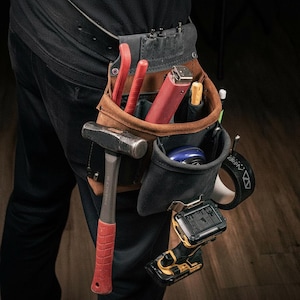 May include: A brown leather and black fabric tool pouch worn on a belt, containing a hammer with a red handle, a red utility tool, a yellow-handled tool, a blue circular object, a black roll of tape, and a yellow and black cordless power drill.