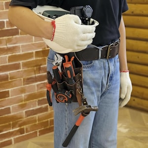 May include: A brown leather tool belt with a selection of tools, including a hammer, pliers, and screwdrivers. The belt is worn with blue jeans and a black shirt. A white hard hat and work gloves are also visible.