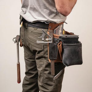 May include: A brown leather tool belt with multiple pockets and compartments, holding various tools including a hammer, square, and measuring tools. The belt is worn by a person wearing olive green pants and a gray t-shirt.