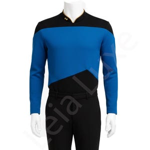 Op de afbeelding: Een blauw en zwart uniformtop met lange mouwen en een zwarte broek. De top heeft een zwarte pas en kraag met gouden accenten. Het blauwe gedeelte wordt verdeeld door een zwarte diagonale lijn. De broek is zwart en recht.