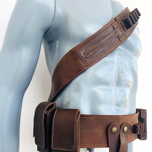May include: A brown leather bandolier and utility belt set featuring multiple pouches, loops for small cylindrical items, and brass-colored metal snap fasteners. The gear is worn across the torso and around the waist, constructed with visible stitching along the edges of the leather components.