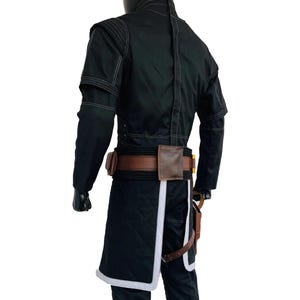 Op de afbeelding: Een zwart jack met lange mouwen, witte stiksels en een hoge kraag, gedragen over een zwarte broek. Een bruine utility-riem met tasjes en een holster zit om de taille. Het jack heeft een gewatteerd zwart paneel met een witte rand aan de zoom.
