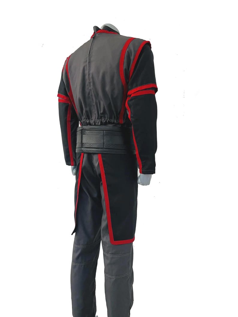 K&ouml;nnte beinhalten: Ein Kost&uuml;m bestehend aus einer anthrazitfarbenen und schwarzen Jacke mit rotem Besatz, langen &Auml;rmeln und einem Stehkragen. Das Outfit umfasst einen breiten schwarzen G&uuml;rtel und eine passende anthrazitfarbene Hose mit sichtbaren N&auml;hten sowie eine Tunika.