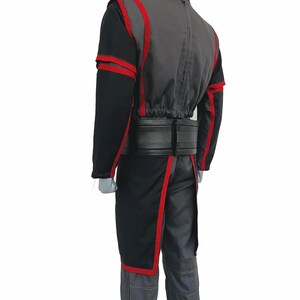 K&ouml;nnte beinhalten: Ein Ganzk&ouml;rperkost&uuml;m bestehend aus einer dunkelgrauen und schwarzen Jacke mit leuchtend roten Zierstreifen. Die Jacke hat einen hohen Kragen, lange &Auml;rmel und einen schwarzen G&uuml;rtel in der Taille. Die passende Hose ist dunkelgrau mit sichtbaren N&auml;hten.