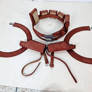 May include: A brown leather utility belt featuring multiple rectangular pouches and a central metal buckle, accompanied by two matching curved leather shoulder holsters with adjustable straps and metallic hardware attachments.