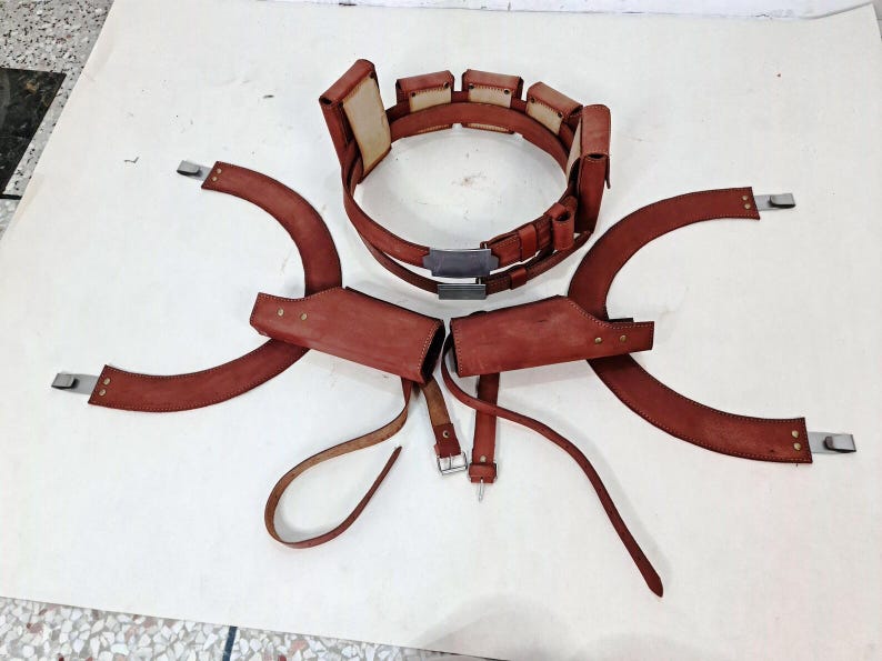 May include: A brown leather utility belt featuring multiple rectangular pouches and a central metal buckle, accompanied by two matching curved leather shoulder holsters with adjustable straps and metallic hardware attachments.